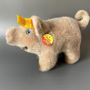 Op de afbeelding: Een pluchen varken met een lichtbruin lichaam, blauwe ogen en een gekrulde staart. Het varken heeft een roze snuit en oren. Een geel label is aan zijn oor bevestigd en een rond label met het woord "Piggy" is aan zijn zijkant bevestigd.