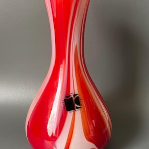 May include: A Murano glass vase with a teardrop shape, featuring a vibrant red and white swirl pattern. The vase has a flared top and a label that reads "Moretti Murano Italia". The glass is glossy and reflects light.