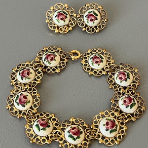 May include: A vintage jewelry set featuring a bracelet and matching earrings. The bracelet and earrings have a gold-tone filigree design. Each piece features a small, round, embroidered floral design with red and green accents on a white background.