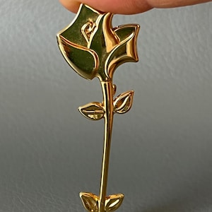 Könnte beinhalten: Eine goldfarbene Rosenbrosche mit detailliertem Design. Die Brosche hat einen langen Stiel mit Blättern und eine voll erblühte Rose. Die Rose wird zwischen zwei Fingern vor einem grauen Hintergrund gehalten. Die Brosche besteht aus glänzendem, reflektierendem Material.
