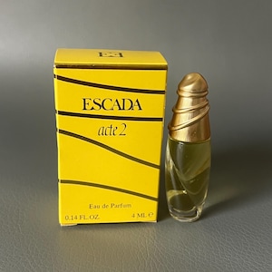 Puede incluir: Una caja amarilla con el texto "ESCADA acte 2" y "Eau de Parfum" con un pequeño frasco de perfume de vidrio transparente con una tapa dorada.