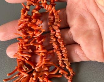May include: A coral necklace with a mix of small, rounded beads and larger, branch-like pieces. The necklace is a vibrant orange-red color and has a gold-colored clasp. The coral is displayed against a neutral background.