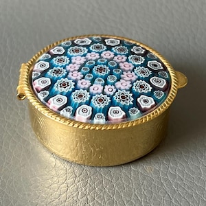 Könnte beinhalten: Eine kleine, runde, goldfarbene Schmuckdose mit dekorativem Deckel. Der Deckel zeigt ein Mosaikmuster aus kleinen, farbenfrohen Blumenmustern in Blau-, Rosa- und Weißtönen. Die Dose hat eine Klappöffnung und einen strukturierten Goldrand.