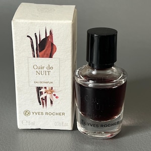 Pode incluir: Um pequeno frasco de perfume com tampa preta ao lado de uma caixa branca. O frasco contém um líquido escuro e tem o texto "Cuir de Nuit". A caixa tem o mesmo texto e um desenho floral.