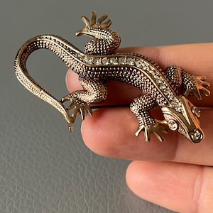 May include: A bronze-toned lizard brooch with a textured body and sparkling clear rhinestones. The lizard has detailed legs, a curled tail, and a decorative head. The brooch is a unique accessory.