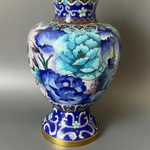 May include: A decorative cloisonné vase with a gold rim and base. The vase features a vibrant floral design in shades of blue, turquoise, and white. The vase is approximately 8 inches tall.