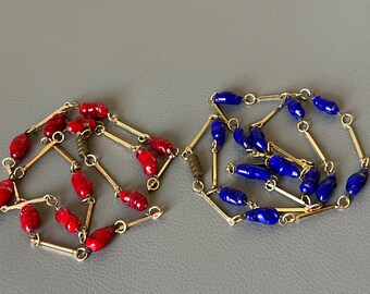 May include: Two necklaces with gold-colored links and beads. One necklace features red beads, the other blue. The beads are oblong and separated by the gold links. The necklaces are displayed on a gray surface.