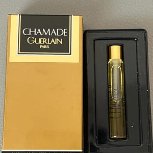 May include: A gold-coloured box and a black tray holding a small perfume vial. The box has the text "CHAMADE GUERLAIN PARIS" in black and gold. The vial contains a light amber liquid. The text "PARFUM DE TOILETTE 2 ml .07 FL.OZ." is printed on the box.