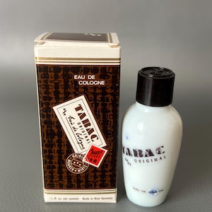 May include: A vintage bottle of Tabac Original Eau de Cologne and its original box. The box is brown with a repeating pattern and the bottle is white with a black cap. The text "TABAC ORIGINAL" is visible on the bottle.