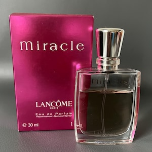 May include: A perfume bottle and box set. The box is a deep pink with the word "miracle" in white. The perfume bottle is clear with a silver cap and contains a light brown liquid. The box reads "Lancôme Paris Eau de Parfum Vaporisateur-Natural Spray e 30 ml".