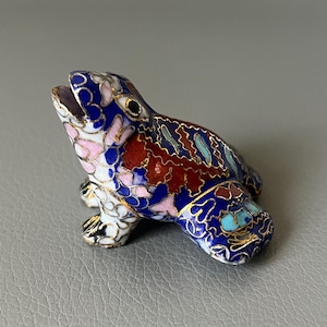 Pode incluir: Estatueta decorativa de sapo com um design cloisonné detalhado. O sapo apresenta uma paleta de cores vibrantes de azul, vermelho, branco, rosa e dourado. Um item decorativo único.