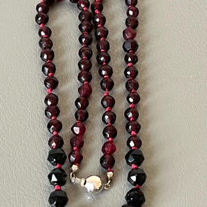 May include: A gemstone necklace with alternating dark red and black faceted beads, strung on a red cord. The necklace has a silver clasp. The beads are cut to reflect light. The necklace is approximately 45 cm long.