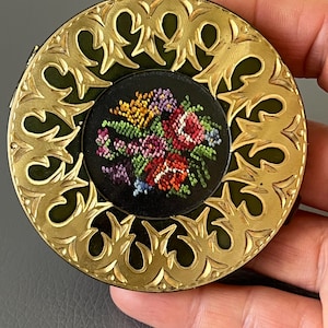 Peut inclure: Un compact rond de couleur or avec un fond vert et un motif floral brodé de fils multicolores.