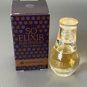 Könnte beinhalten: Eine kleine Flasche Yves Rocher So Elixir Bois Sensuel Parfüm neben der passenden Schachtel. Die Flasche ist klar und enthält eine goldfarbene Flüssigkeit. Die Schachtel ist dunkelviolett mit goldenen Akzenten.