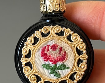 Op de afbeelding: Een kleine, zwarte parfumfles met een gouden filigraan deksel. De fles heeft een cirkelvormig geborduurd ontwerp van een roze roos met groene bladeren, omlijst door een gouden filigraan rand. De fles wordt vastgehouden door de vingers van een persoon.
