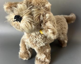 May include: A plush, light brown dog toy with a fluffy texture. The toy has black embroidered eyes and a yellow collar. The dog is standing on all fours, with its tail slightly curved. The toy is made of soft, fuzzy material.