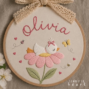 Op de afbeelding: Geborduurde borduurring met de naam "Olivia" in roze draad. Een witte vogel met een roze strik rust op een roze bloem. Een lieveheersbeestje, vlinder en bij zijn ook geborduurd. De ring is versierd met een beige kanten strik.