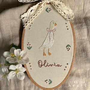 May include: Oval embroidered wall hanging featuring a white goose with a pink bow, the name "Olivia", and floral accents. The piece is framed in a wooden hoop and adorned with a lace bow at the top.