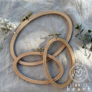 Puede incluir: Tres bastidores de bordado ovalados de madera de diferentes tamaños. Los bastidores son de color marrón claro con herrajes de color latón. La imagen incluye ramitas de flores blancas y hojas verdes. El texto "SEWN TO HEART" es visible en la esquina inferior derecha.