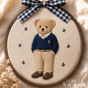 Könnte beinhalten: Ein runder Stickrahmen mit einem beigen Teddybären, der einen marineblauen Pullover, ein weißes Hemd und eine beige Hose trägt. Der Bär hat braune Schuhe und ein kleines goldenes Logo. Oben ist eine marineblau-weiße Vichy-Schleife befestigt. Text: SEWN TO heart.