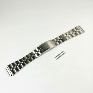 Peut inclure: Un bracelet de montre en acier inoxydable argenté avec un fermoir déployant et deux barrettes à ressort. Le bracelet est composé de maillons rectangulaires et est conçu pour être fixé à un boîtier de montre. Neuf et non utilisé.