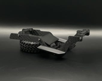 May include: A black radio-controlled vehicle chassis frame constructed from textured material with multiple mounting holes, a central flat plate, and a single black rubber tire with a deep tread pattern attached to the rear assembly.