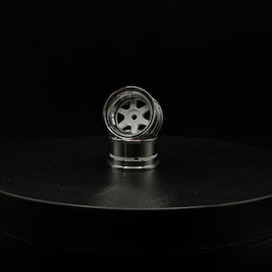 May include: Two miniature chrome-plated model car wheels stacked on top of each other. The wheels have a silver finish with a white, six-spoke design in the center. The wheels are set against a dark background.