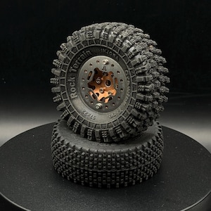 Peut inclure: Deux pneus noirs de voiture RC empilés. Le pneu supérieur a une jante métallique avec un centre de couleur cuivre et les mots "Rock Terrain" et "Injora" sur le pneu. Le pneu inférieur est également noir et présente un motif de bande de roulement similaire.