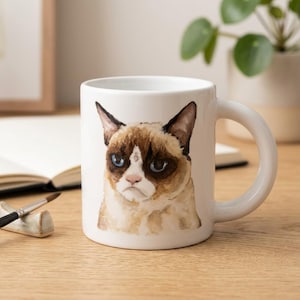 May include: A white ceramic mug featuring a watercolour illustration of a cat with brown and cream fur, blue eyes, and a stern facial expression. The cat is centred on the front of the mug, which has a smooth finish and a curved handle on the right side.