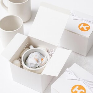 May include: A set of white ceramic mugs, with one mug nestled inside an open white cardboard gift box filled with white paper shred. Two additional closed white gift boxes are tied with white string and feature an orange circular logo with the letters HC.