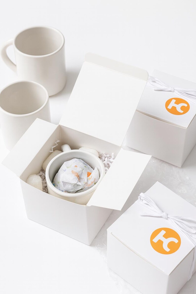 May include: A set of white ceramic mugs, with one mug nestled inside an open white cardboard gift box filled with white paper shred. Two additional closed white gift boxes are tied with white string and feature an orange circular logo with the letters HC.