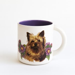 Puede incluir: Taza de cerámica blanca con interior morado, con una ilustración detallada de un perro marrón y canela con flores moradas. La taza tiene un asa blanca y está sobre un fondo blanco.