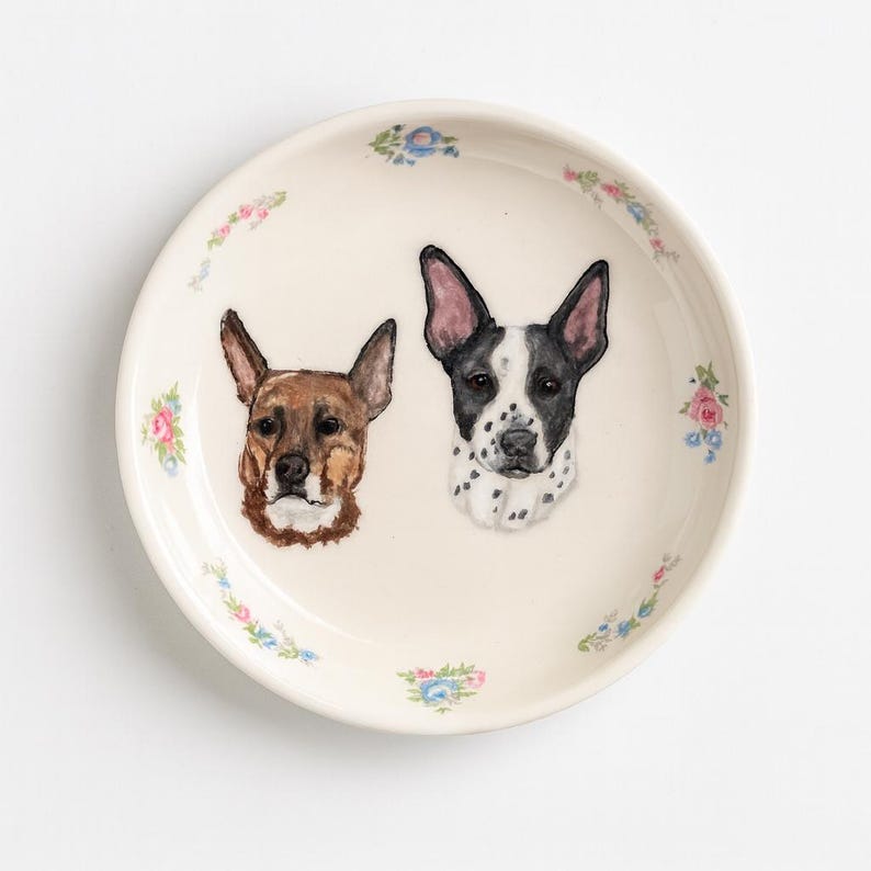 Puede incluir: Un plato redondo de cer&aacute;mica blanquecina con retratos pintados a mano de dos perros. Uno es marr&oacute;n, el otro blanco y negro. El plato est&aacute; decorado con peque&ntilde;os motivos florales en rosa, azul y verde.