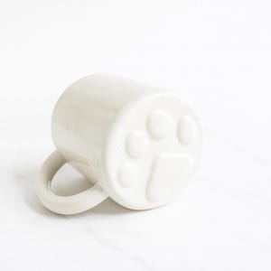 May include: A glossy white ceramic mug featuring a raised, embossed paw print design on the bottom exterior. The mug has a smooth, cylindrical body and a curved handle extending from the side, resting on its side to display the paw detail.