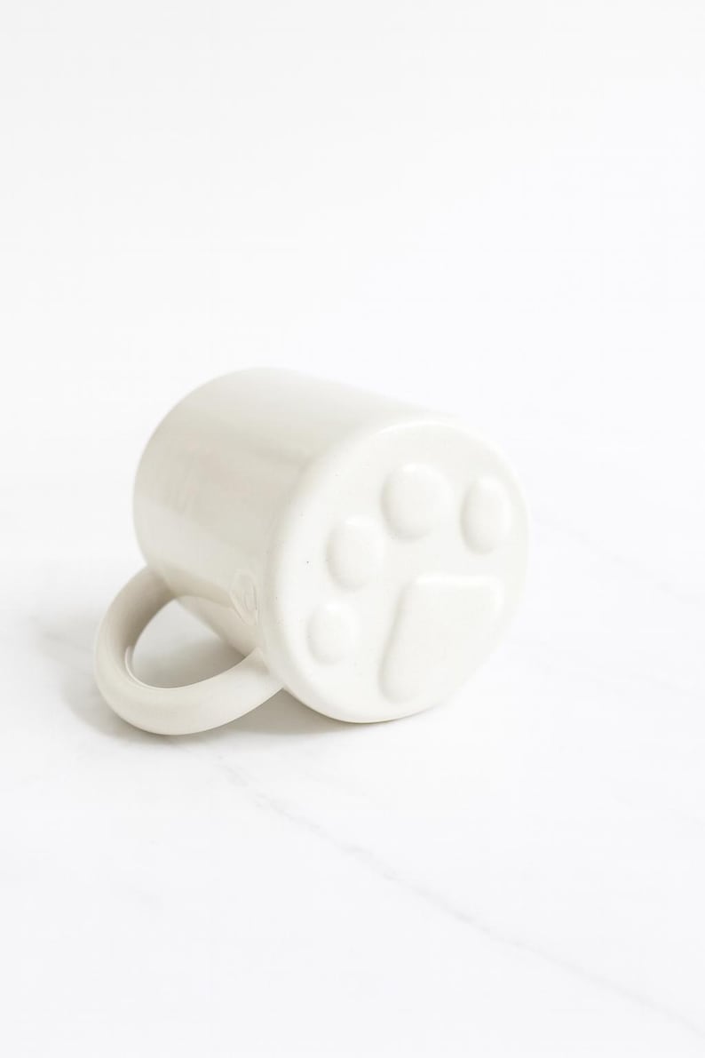 May include: A glossy white ceramic mug featuring a raised, embossed paw print design on the bottom exterior. The mug has a smooth, cylindrical body and a curved handle extending from the side, resting on its side to display the paw detail.