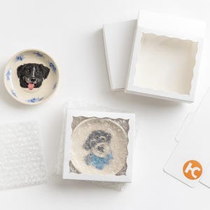 Puede incluir: Dos platos de cer&aacute;mica con retratos de perros, uno con un perro blanco y negro con la lengua fuera, y otro con una bufanda azul. Los platos est&aacute;n empaquetados en cajas blancas con pl&aacute;stico de burbujas.