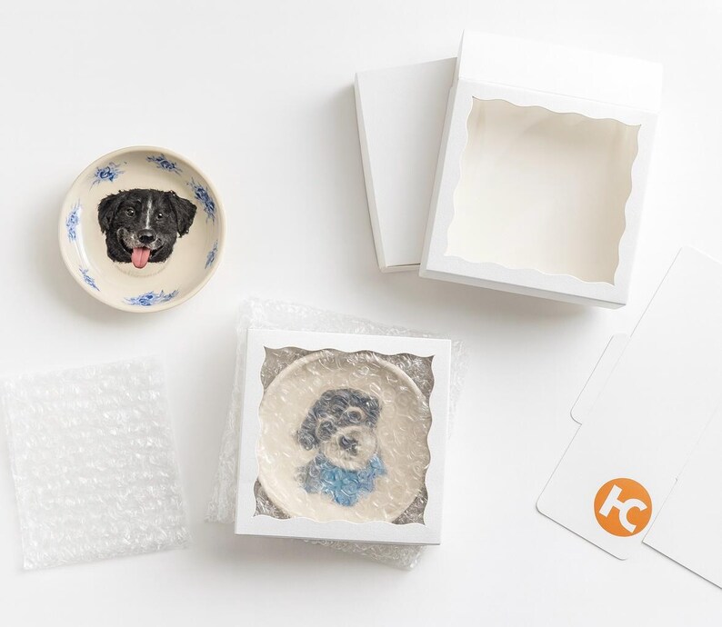 Puede incluir: Dos platos de cer&aacute;mica con retratos de perros, uno con un perro blanco y negro con la lengua fuera, y otro con una bufanda azul. Los platos est&aacute;n empaquetados en cajas blancas con pl&aacute;stico de burbujas.