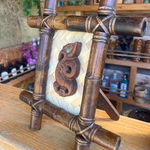 Könnte beinhalten: Ein handgefertigter Bilderrahmen aus Bambus mit einem gewebten Mittelteil. Der Rahmen zeigt eine geschnitzte Holzfigur mit türkisfarbenem Akzent. Der Rahmen wird mit gewebtem Bindfaden zusammengehalten und hat ein natürliches, rustikales Aussehen.