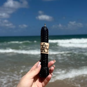 May include: A black honeycomb candle with a brown paper label that says "A5G" in black lettering. The candle is held in a hand against a background of a beach and ocean.