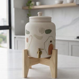 Peut inclure: Un distributeur de boissons en céramique crème avec un couvercle, des motifs peints abstraits et un robinet en laiton. Il repose sur un support en bois clair. Le distributeur est dans une cuisine.