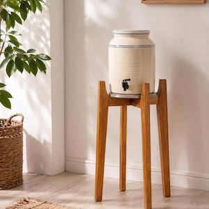 May include: A cream ceramic water dispenser with blue stripes, on a wooden stand. The dispenser has a black tap and a lid. The stand has four angled legs. The setting is a bright room with a plant and a woven basket.