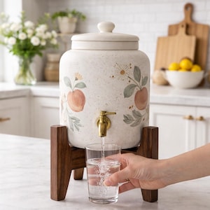 Peut inclure: Un distributeur d'eau en céramique crème avec un couvercle, orné d'illustrations de pêches et de fleurs. Il est équipé d'un robinet en laiton et repose sur un support en bois foncé. Un verre est rempli d'eau.