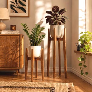 May include: Two wooden plant stands with four legs each, holding potted plants. One stand is shorter with a speckled white pot, and the other is taller with a ribbed white pot. The stands are made of light brown wood with a smooth finish.