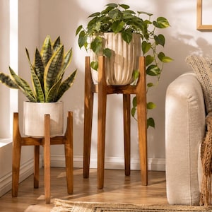 Peut inclure: Deux supports de plantes en bois à quatre pieds chacun, accueillant deux plantes en pot différentes. Le support le plus court porte un pot blanc texturé avec une sansevieria, tandis que le plus haut porte un pot crème cannelé avec un pothos.