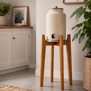 May include: A cream-colored ceramic beverage dispenser with vertical ribbed detailing, a wooden lid, and a black plastic spigot. The dispenser rests on a tall, four-legged wooden stand with a natural brown finish and a circular metal base support.