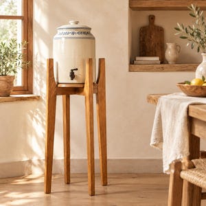 May include: A cylindrical ceramic water dispenser with a white glaze and a blue floral pattern around the top rim. It features a black spigot and a matching lid, resting on a tall, four-legged wooden stand with a natural light brown finish.