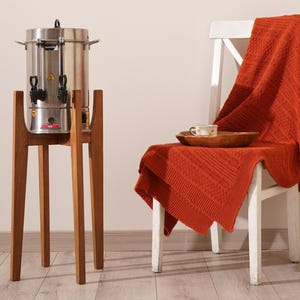 May include: A stainless steel coffee server with two spouts sits on a wooden stand. A red blanket is draped over a white chair with a wooden tray and a cup on top.