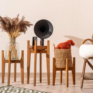 Peut inclure: Trois supports de plantes en bois de différentes hauteurs. L'un contient un vase en verre avec des herbes séchées, un autre un gramophone vintage et le troisième un panier en osier avec un pull orange. Une chaise en osier est à droite.