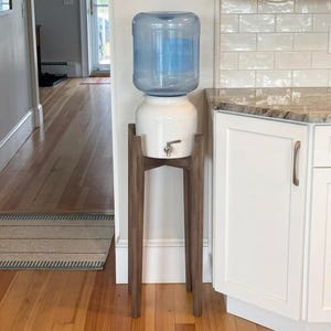 May include: A water dispenser featuring a large, clear plastic jug resting on a white ceramic crock base. The unit is supported by a tall, four-legged wooden stand with a dark brown finish and includes a silver-toned metal spigot for dispensing.