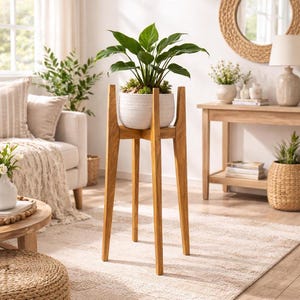 May include: A tall, wooden plant stand with a white ceramic pot holding a green leafy plant. The stand has a tripod base and a square top to hold the pot. The stand is made of light-colored wood and is in a living room setting.